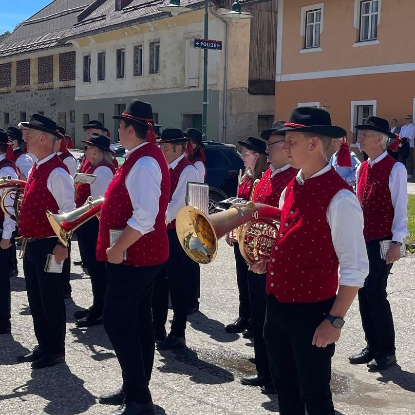 Eine Blaskapelle spielt draußen, Musiker in roten Westen halten Blechblasinstrumente. Sie stehen vor Gebäuden mit einigen Fenstern und einem Straßenschild.