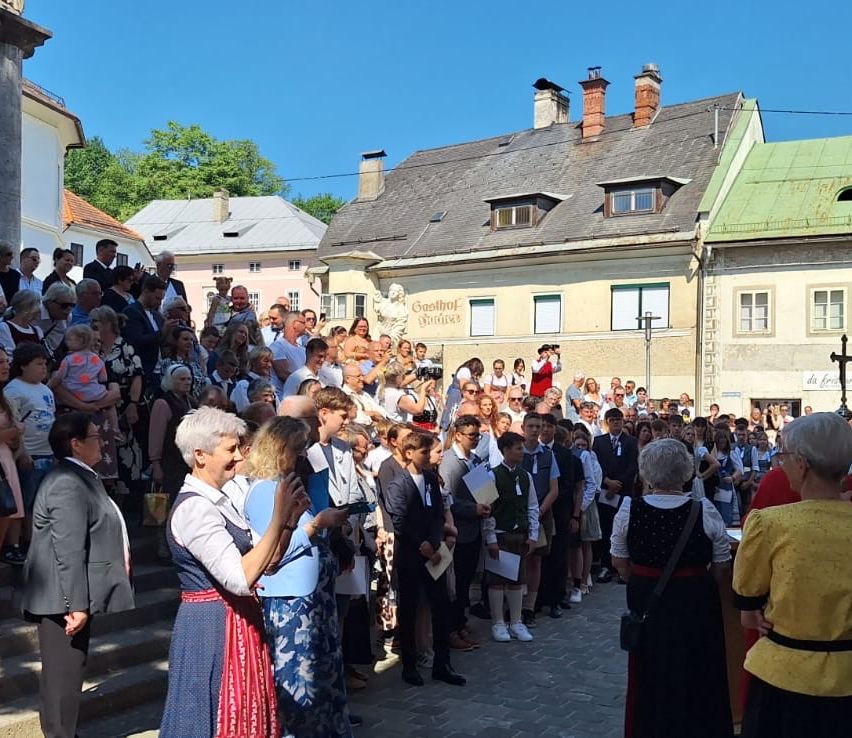 Eine große Menschenmenge versammelt sich auf einem Stadtplatz, einige in traditioneller Kleidung, andere in formeller Kleidung. Ein Schild am Gebäude lautet Goethehof.