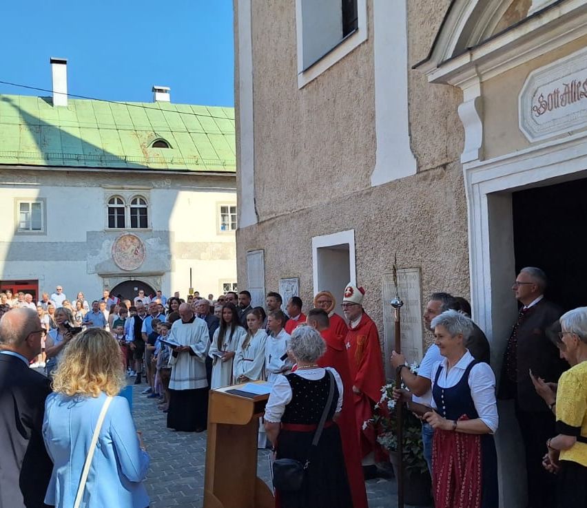 Eine große Versammlung von Menschen, einschließlich Geistlicher und Gemeindemitglieder, findet vor einer Kirche statt. Einige Personen tragen traditionelle Kleidung.