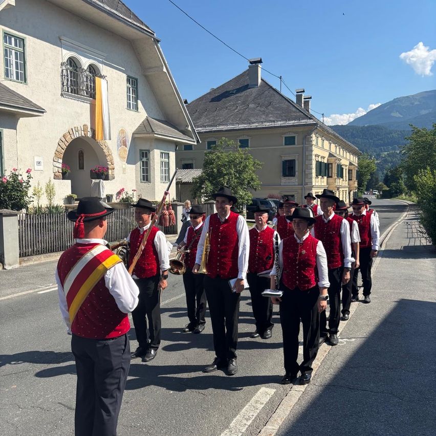 Eine Blaskapelle, gekleidet in rote Westen und schwarze Hosen, tritt auf einer Straße auf, mit Gebäuden und Bergen im Hintergrund.