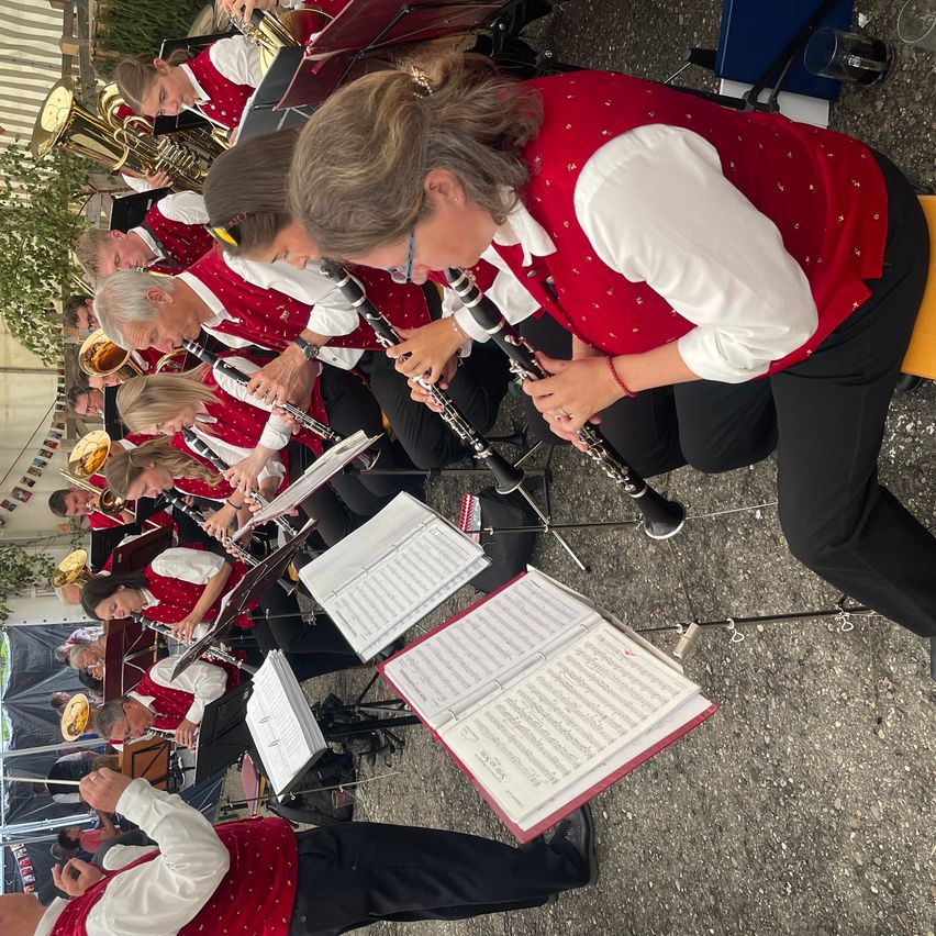 Eine Gruppe von Musikern in roten Westen spielt auf der Bühne Instrumente. Eine Frau vorne spielt Klarinette. Vor ihnen stehen Musikblätter auf einem Ständer.