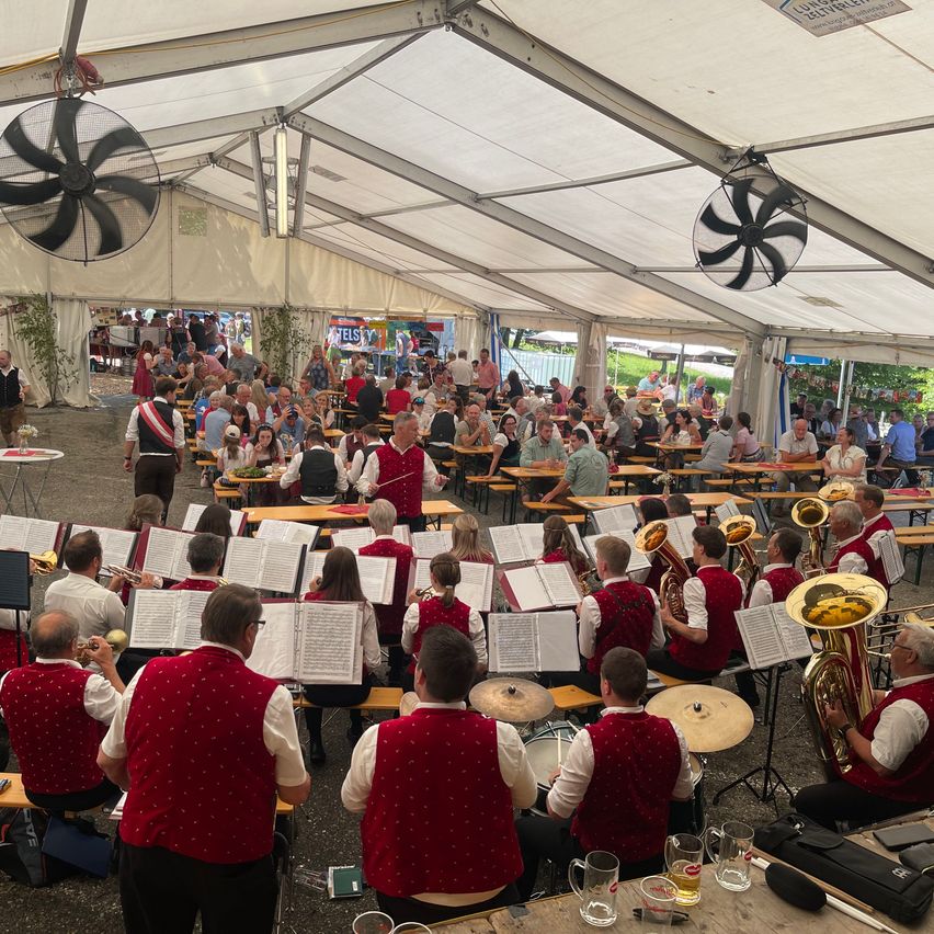 Eine Outdoor-Band spielt für ein Publikum bei einer Veranstaltung unter einem großen Zelt mit Ventilatoren. Die Bandmitglieder sitzen auf Bänken und spielen Instrumente, während viele Menschen zuschauen.