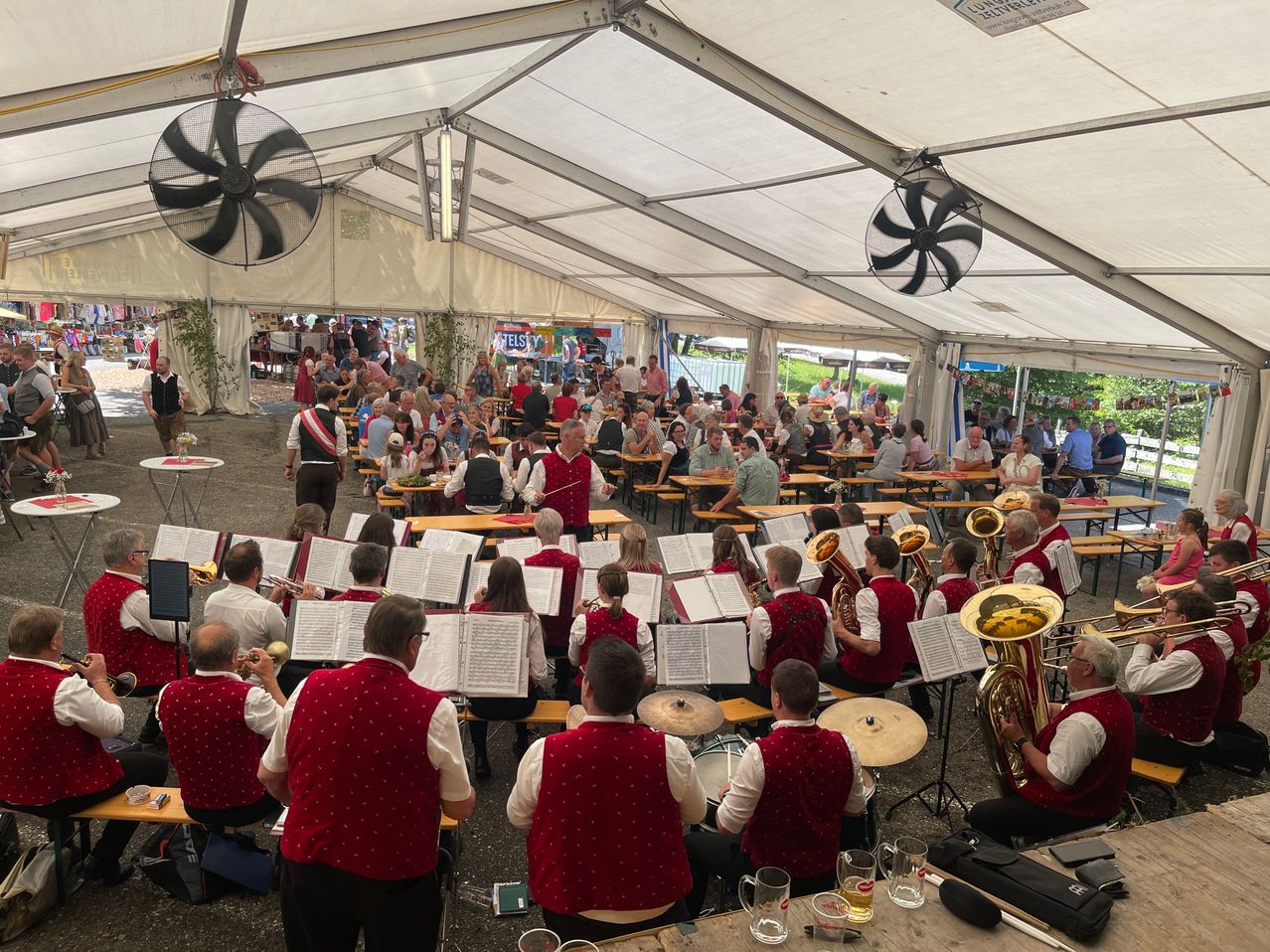 Eine Outdoor-Band spielt für ein Publikum bei einer Veranstaltung unter einem großen Zelt mit Ventilatoren. Die Bandmitglieder sitzen auf Bänken und spielen Instrumente, während viele Menschen zuschauen.