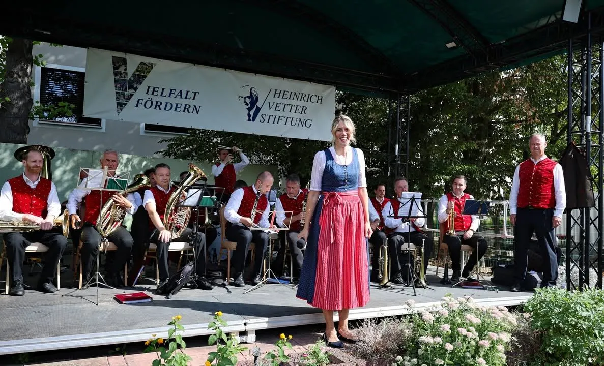 Eine Frau in einem traditionellen Kleid steht auf der Bühne mit einer Band hinter ihr. Die Band spielt verschiedene Instrumente, und die Bühne ist mit Blumen geschmückt. Ein Banner mit Text hängt über ihnen.