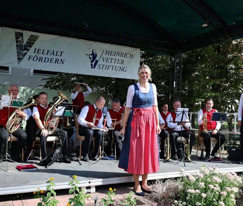 Eine Frau in einem traditionellen Kleid steht auf der Bühne mit einer Band hinter ihr. Die Band spielt verschiedene Instrumente, und die Bühne ist mit Blumen geschmückt. Ein Banner mit Text hängt über ihnen.