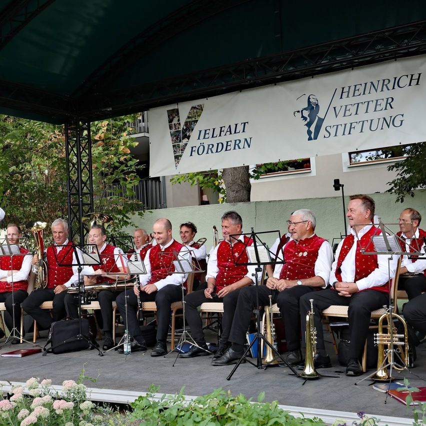 Eine Gruppe von Musikern in roten Westen und schwarzen Hosen sitzt auf Stühlen auf einer Bühne mit Musikinstrumenten und einem Banner dahinter.