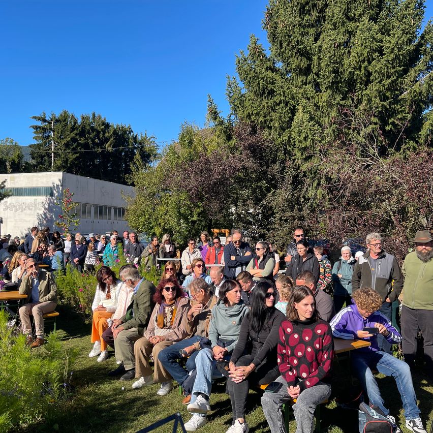 Eine große Gruppe von Menschen sitzt auf Bänken in einem Park, einige stehen und andere sitzen an Tischen. Sie sind von Grün und Bäumen umgeben, und ein Gebäude ist im Hintergrund zu sehen.