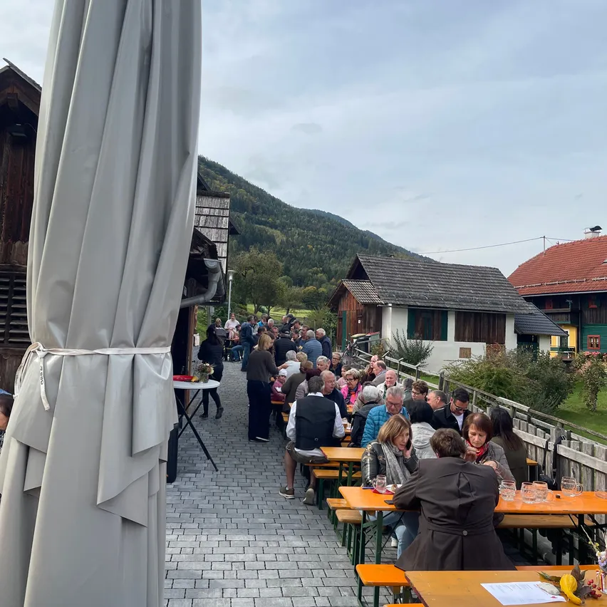Eine Gruppe von Menschen sitzt an Tischen in einem Restaurant im Freien. Das Restaurant ist von Häusern und Bergen umgeben.