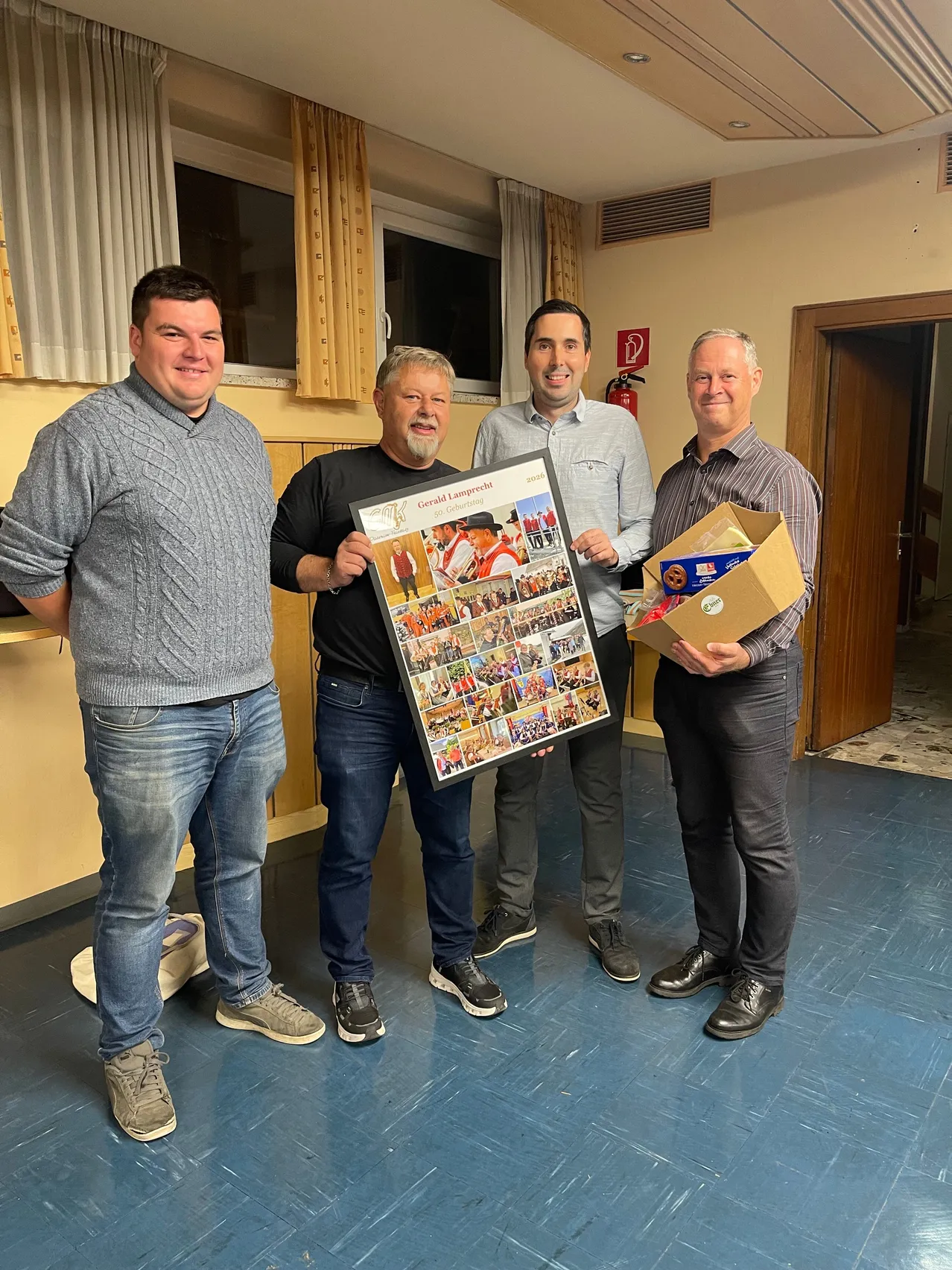 Vier Männer stehen in einem Raum, lächeln und posieren für ein Foto. Ein Mann hält ein großes Poster mit Bildern von Menschen und ein Feuerlöscher ist an der Wand sichtbar.