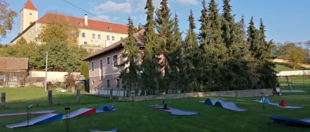 Ein Freiluftbereich mit einem grünen Rasen hat einen Minigolfplatz mit verschiedenen Rampen und Abhängen. Bäume umgeben den Bereich und es gibt ein Gebäude im Hintergrund.