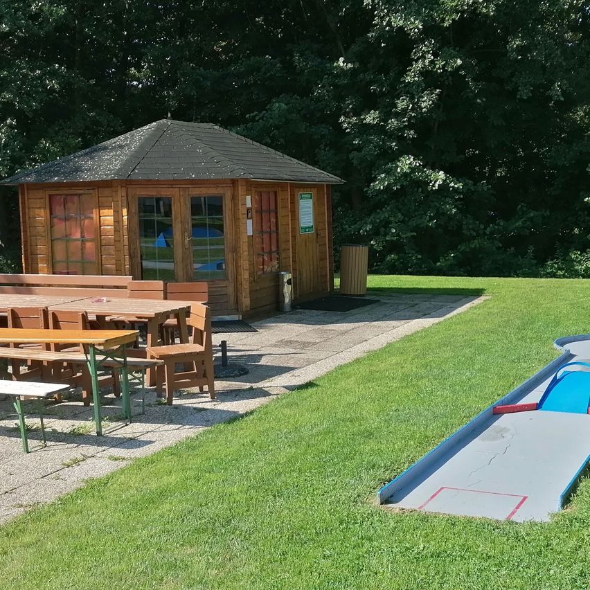 Ein kleines Holzhaus mit Glasfenstern und einem Mülleimer steht auf einer Rasenfläche. Ein Tisch mit Stühlen und eine Bank sind in der Nähe platziert. Ein Minigolfplatz befindet sich vor dem Haus.