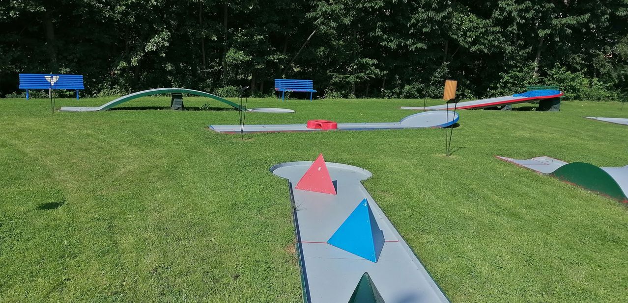 Ein Golfplatz mit grünem Rasen und Hindernissen, bestehend aus einer blauen Bank, einer roten und blauen Pyramide sowie geometrischen Formen auf dem Boden.