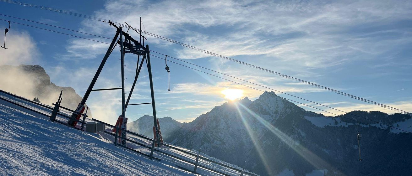 Eine Skiliftstation mit einer Gondel, die sich unter der hellen Sonne den verschneiten Berg hinauf bewegt. Die Berge sind mit Schnee bedeckt.