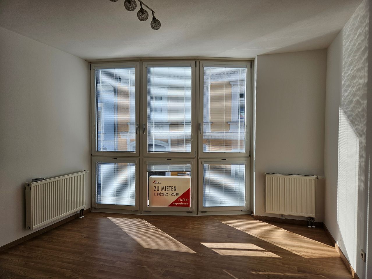 Ein leerer Raum mit Holzboden, weißen Wänden und großen Fenstern mit Jalousien. Der Raum hat zwei weiße Heizkörper auf beiden Seiten und ein Schild am Fenster, das 'Zu Mieten' besagt.