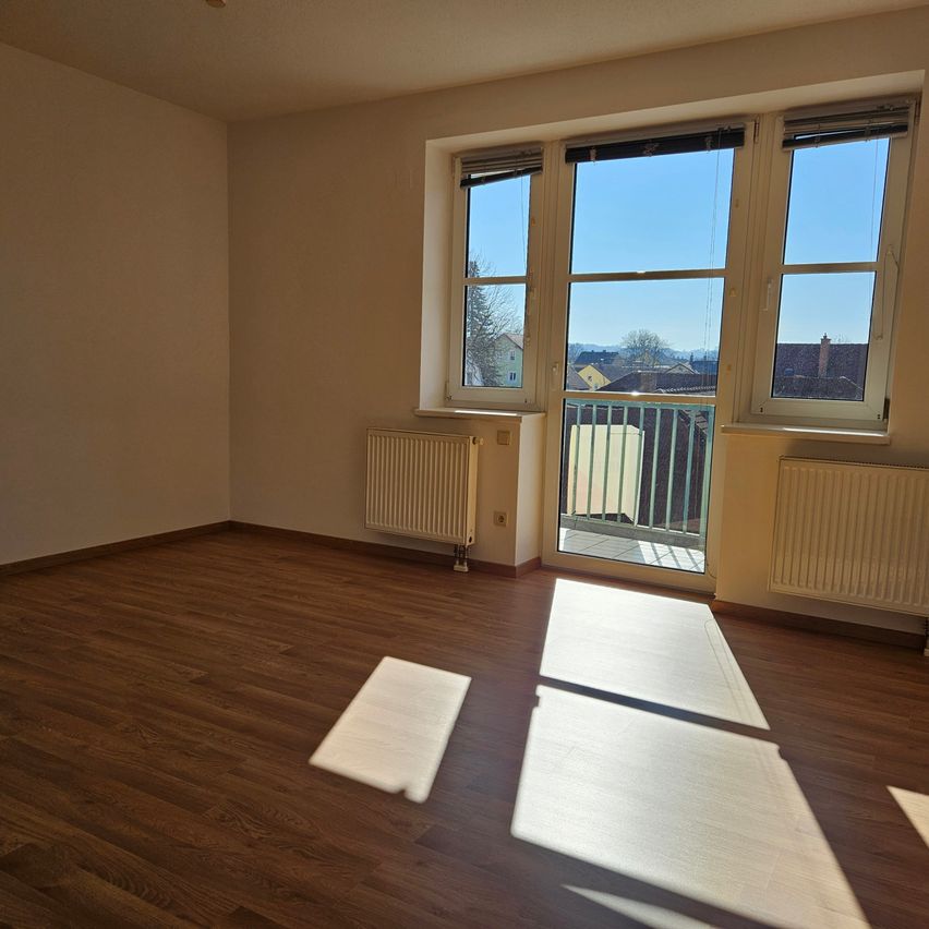 Leerer Raum mit Holzböden und weißen Wänden, zwei große Fenster mit weißen Rahmen und offenen Fensterläden, eines mit Balkon und Geländer, und zwei weiße Heizkörper an den Wänden.