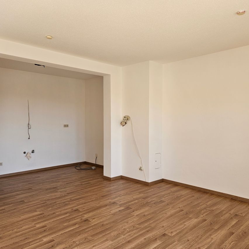 Ein leerer Raum mit weißen Wänden und einem Holzboden, an der Wand befinden sich einige Steckdosen und ein Kabel hängt an der Wand.