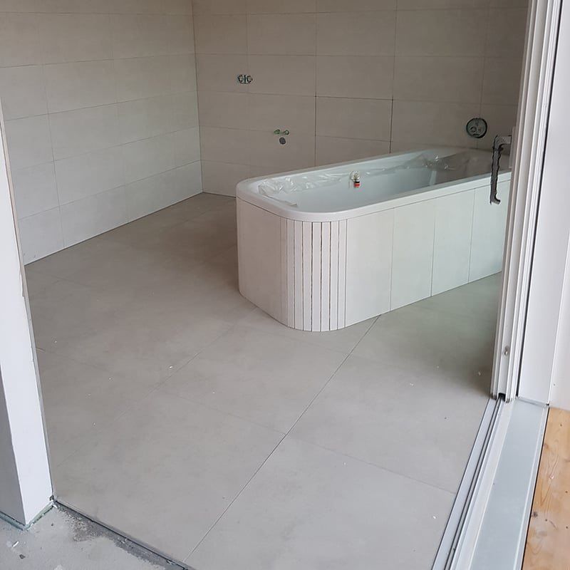 Ein leeres Badezimmer mit weißen gefliesten Wänden und Boden. Ein weißer Badewannenrand ist teilweise sichtbar in der Ecke.