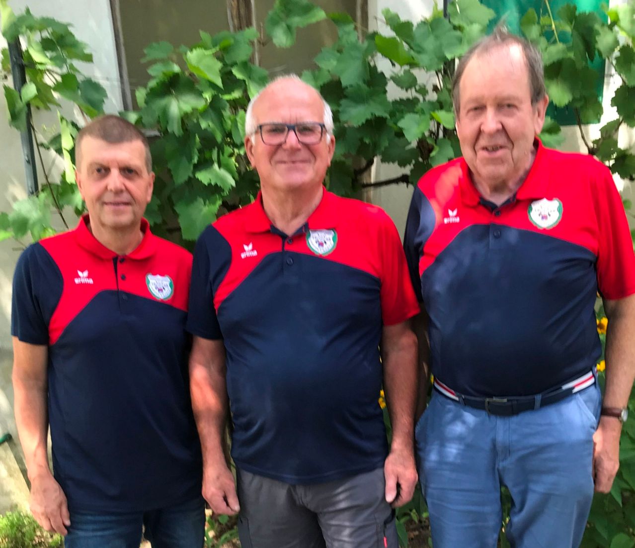 Three men in matching red and blue polo shirts with a logo stand together in front of a building with greenery.