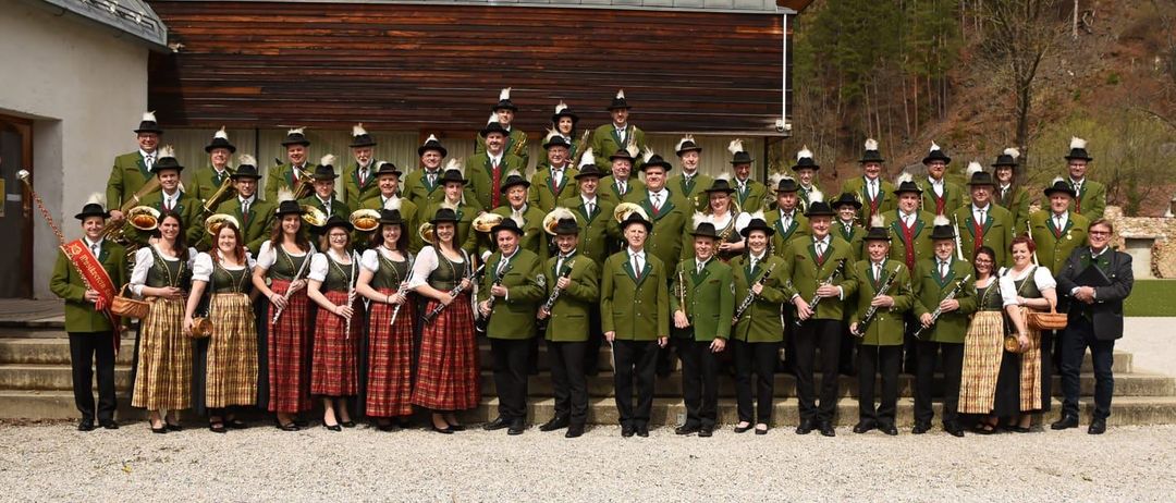 Ein Musikorchester steht auf einem Kiesplatz vor einem Gebäude mit einem Berg im Hintergrund. Sie tragen grüne Uniformen, Hüte und halten Musikinstrumente.