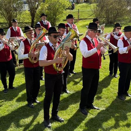 Eine Gruppe von Musikern in roten Westen und schwarzen Hüten spielt auf einer Rasenfläche und benutzt Instrumente wie Trompeten, Tuben und Trommeln.