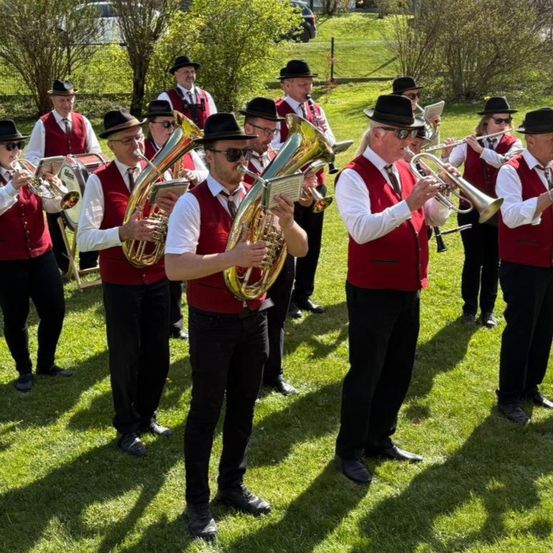 Eine Gruppe von Musikern in roten Westen und schwarzen Hüten spielt auf einer Rasenfläche und benutzt Instrumente wie Trompeten, Tuben und Trommeln.