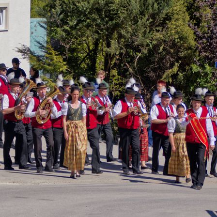 Eine Blaskapelle spielt auf der Straße. Sie tragen rote Westen und spielen Blechblasinstrumente. Eine Frau in einem bunten Rock steht vorne.