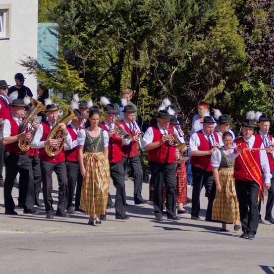 Eine Blaskapelle spielt auf der Straße. Sie tragen rote Westen und spielen Blechblasinstrumente. Eine Frau in einem bunten Rock steht vorne.
