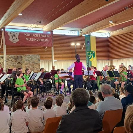 Ein Musikensemble tritt auf der Bühne in einem Raum mit Holzwänden, einer Steinwand und einem Banner mit der Aufschrift Kreuzberger Musikverein auf. Mehrere Kinder sitzen auf Stühlen, während andere stehen und die Aufführung beobachten.