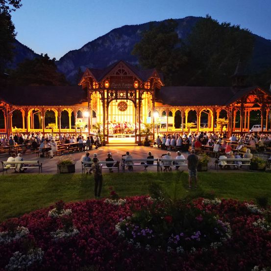 Ein nächtliches Open-Air-Konzert in einem Pavillon mit Blumen, Menschen auf Bänken und einer Bergkette im Hintergrund.