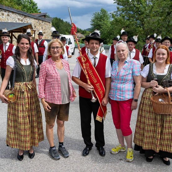 Eine Gruppe von Menschen in traditioneller bayerischer Tracht posiert für ein Foto, ein Mann hält eine Trompete. Dahinter stehen weitere Personen in ähnlicher Kleidung mit Musikinstrumenten.