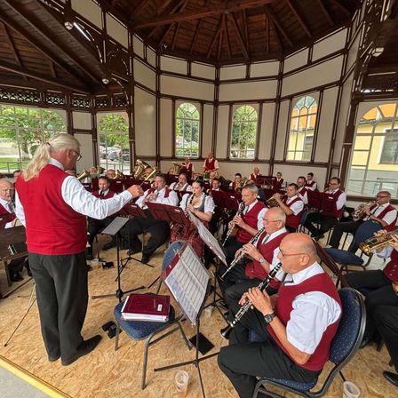 Eine Band probet in einem Pavillon mit roter Weste, hält eine Musikpartitur, während Musiker verschiedene Instrumente spielen, darunter Trompeten und Klarinetten.
