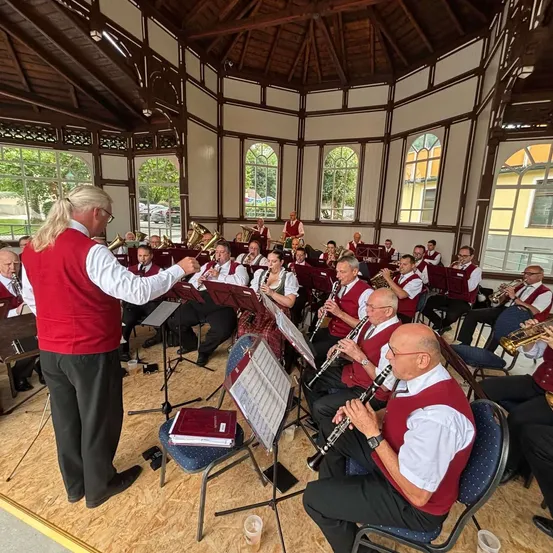 Eine Band probet in einem Pavillon mit roter Weste, hält eine Musikpartitur, während Musiker verschiedene Instrumente spielen, darunter Trompeten und Klarinetten.