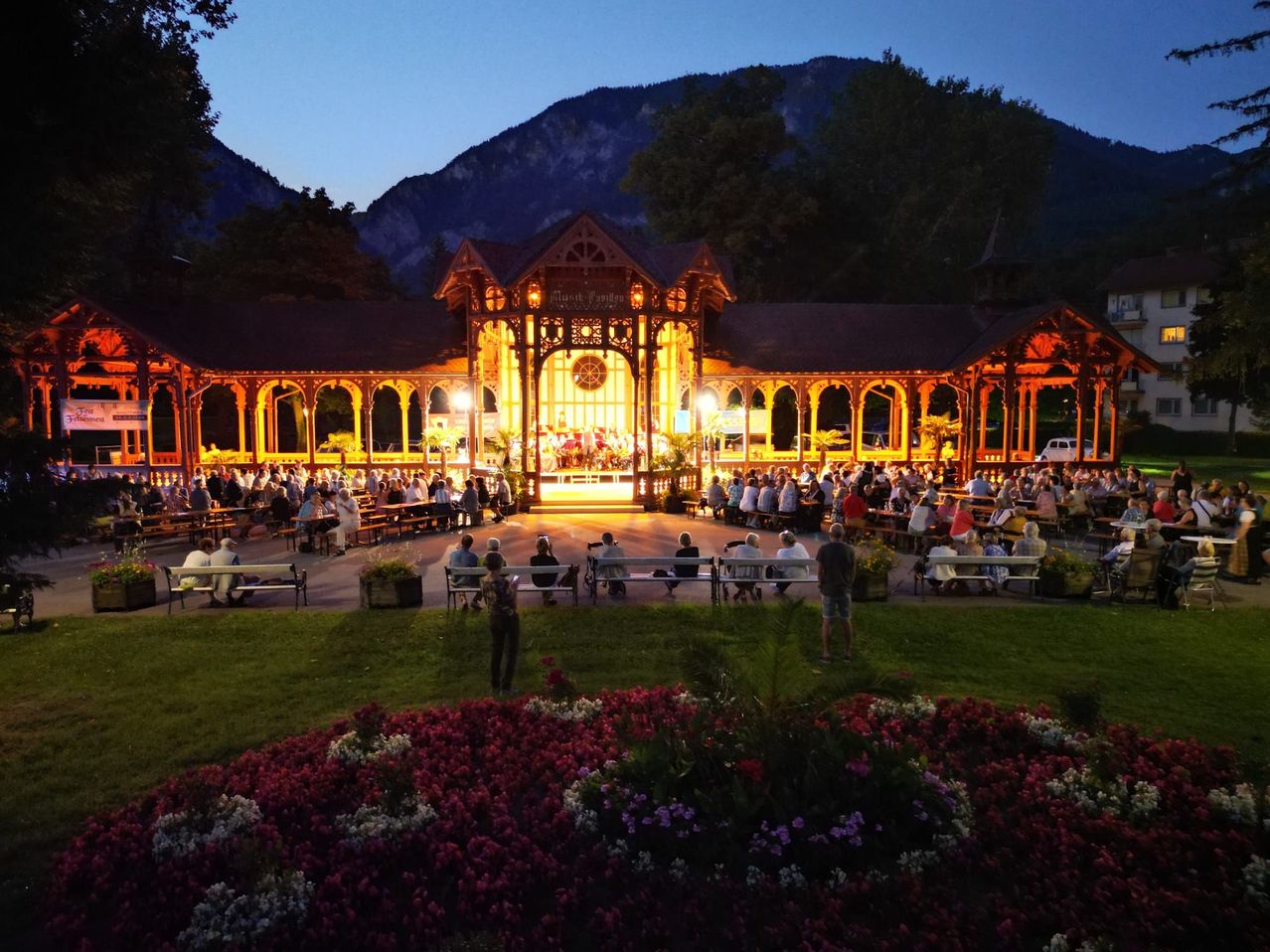 Ein Abendvorstellung in einem Pavillon mit vielen zuschauenden Personen, beleuchtet von warmen Lichtern, umgeben von Blumen. In der Ferne sind Berge sichtbar.