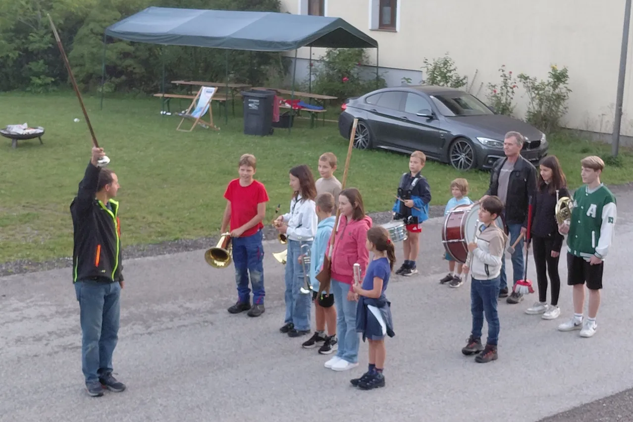 Eine Gruppe von Kindern spielt Instrumente auf einem Rasenstück mit einem Vordach und einem geparkten Auto. Einige Erwachsene sind anwesend, und ein paar Leute stehen in der Nähe.