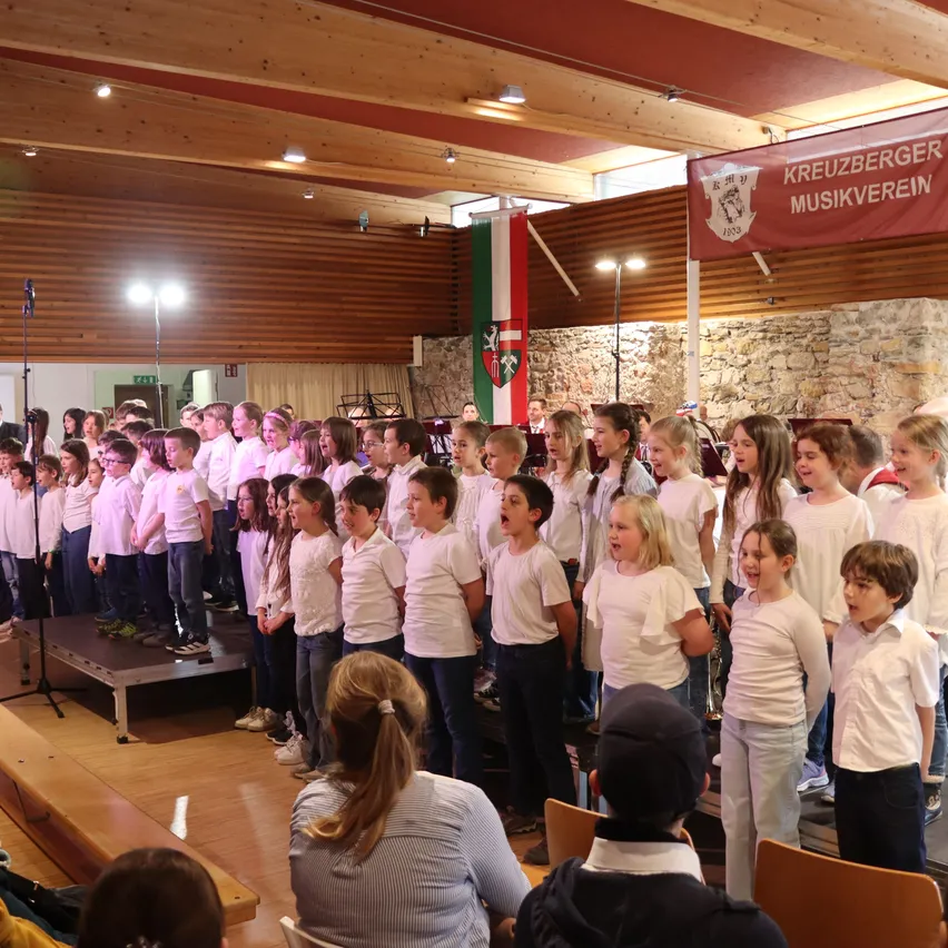 Ein Kinderchor, in Weiß gekleidet, steht auf der Bühne und singt in einem Saal mit Holzwänden und Decke, geschmückt mit einer Fahne und einem Banner. Zuschauer sitzen unten.