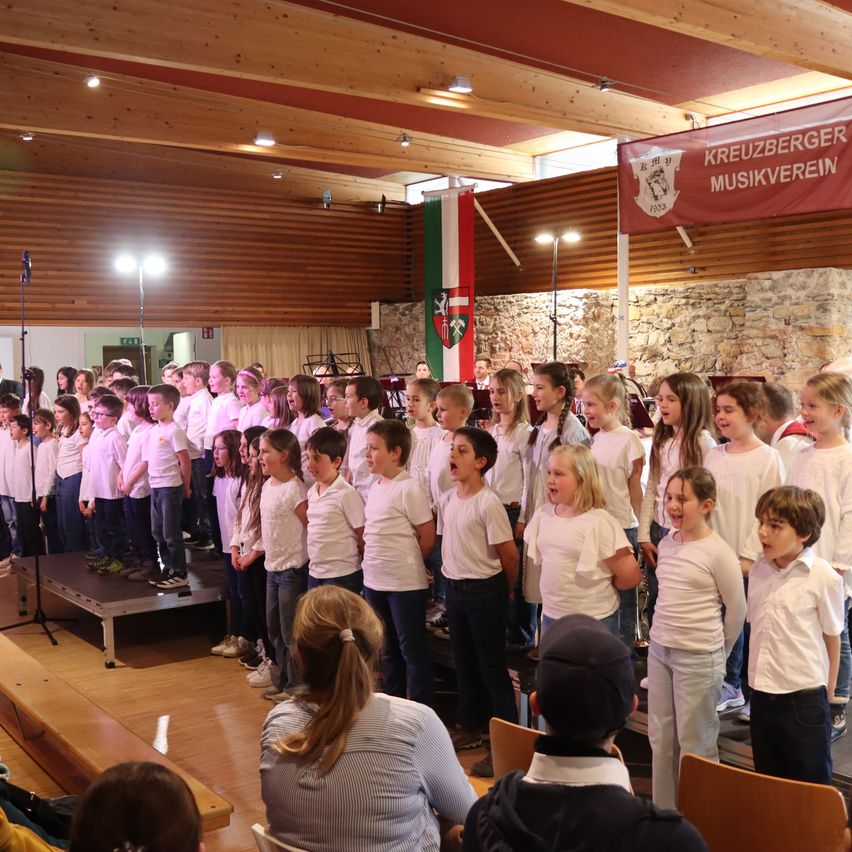 A choir of children, dressed in white, stands on stage singing in a hall with wooden walls and ceiling, adorned with a flag and banner. Audience members sit below.