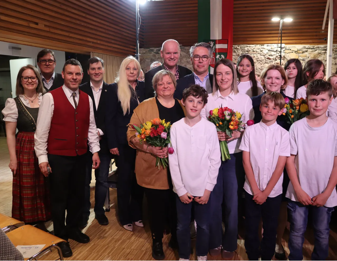 Eine Gruppe von Erwachsenen und Kindern steht in einem Raum zusammen. Sie tragen alle festliche Kleidung. Die Frau in der Mitte hält einen Blumenstrauß. Der Mann auf der linken Seite trägt eine rote Weste. Hinter ihnen ist eine Wand mit einer Fahne.