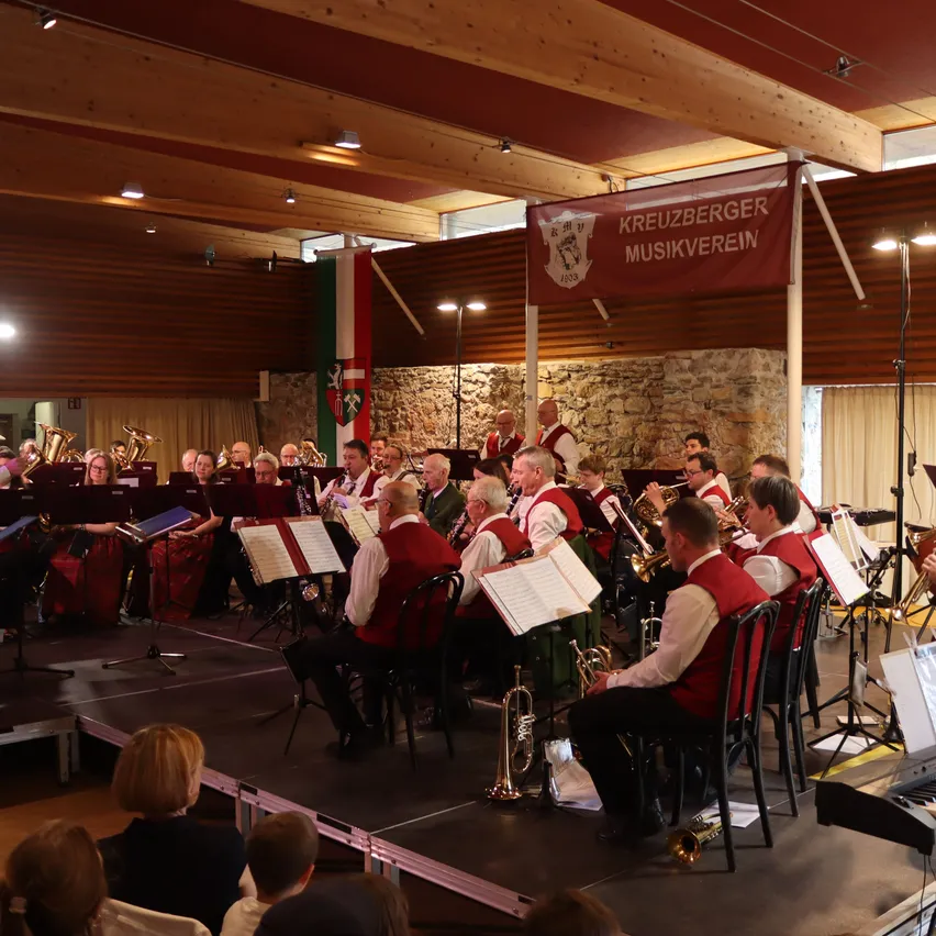 Ein Orchester spielt auf der Bühne mit verschiedenen Musikinstrumenten. Zuschauer sitzen unten und schauen zu. Ein Banner mit dem Text Kreuzberger Musikverein hängt im Hintergrund.