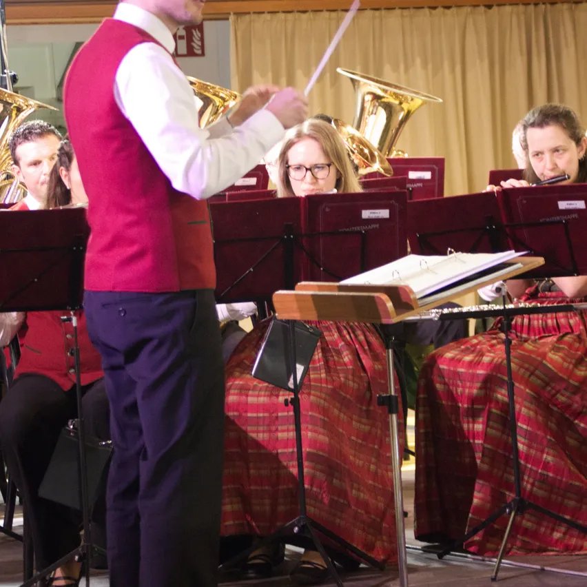 Ein Dirigent steht vor einem Blechbläser-Ensemble in einem Konzertsaal, wobei die Musiker sitzen und ihre Instrumente spielen.