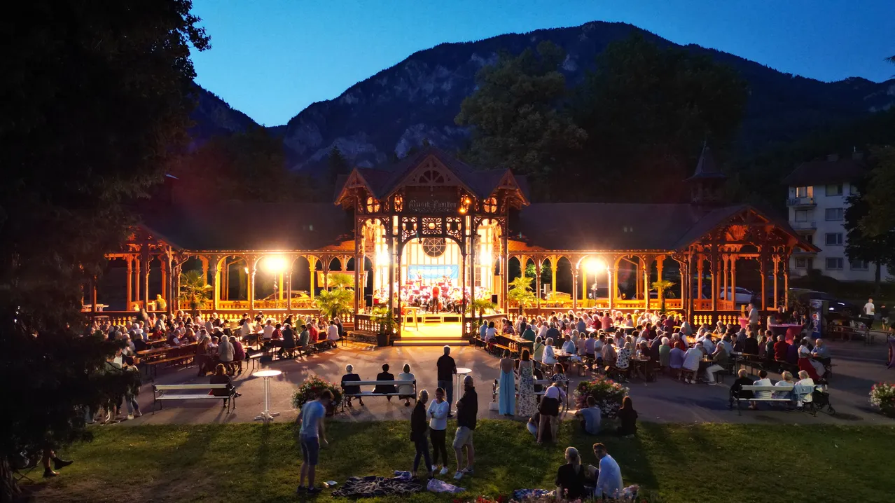 Ein nächtliches Konzert in einem Pavillon im Freien mit einer Menschenmenge, die auf Bänken sitzt und herumsteht, beleuchtet von Lampen. Im Hintergrund befindet sich eine Bühne mit einer auftretenden Band, und Berge sind in der Ferne sichtbar.