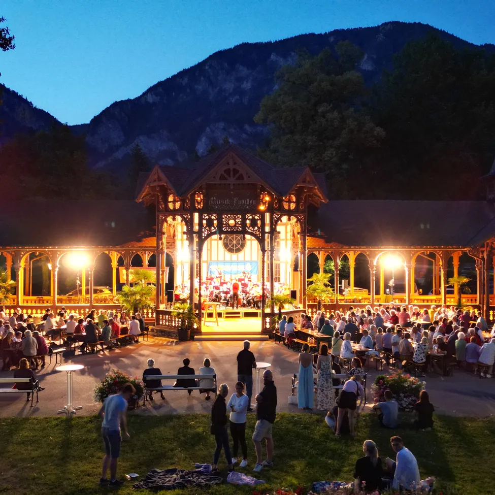 Ein nächtliches Konzert in einem Pavillon im Freien mit einer Menschenmenge, die auf Bänken sitzt und herumsteht, beleuchtet von Lampen. Im Hintergrund befindet sich eine Bühne mit einer auftretenden Band, und Berge sind in der Ferne sichtbar.
