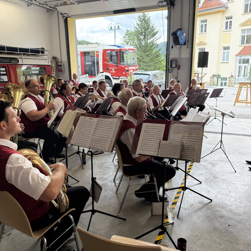 Ein Blasorchester spielt in einer Feuerwache, mit einem roten Feuerwehrauto draußen geparkt.