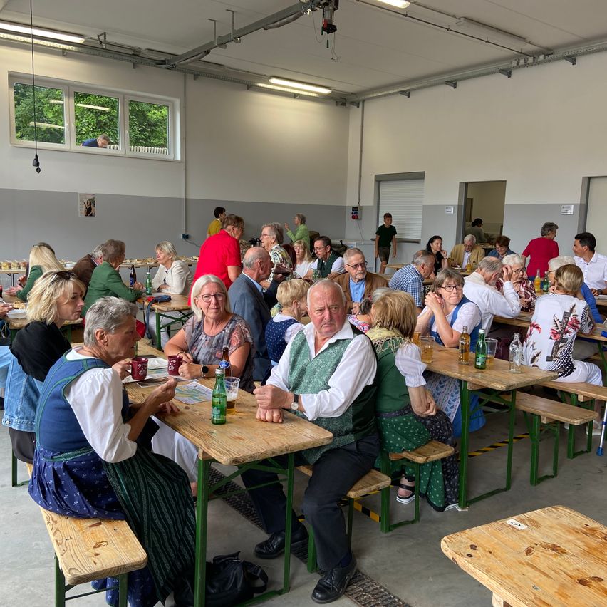 Gäste in Tracht und Anzügen sitzen bei einem Fest im Freien an Holztischen.