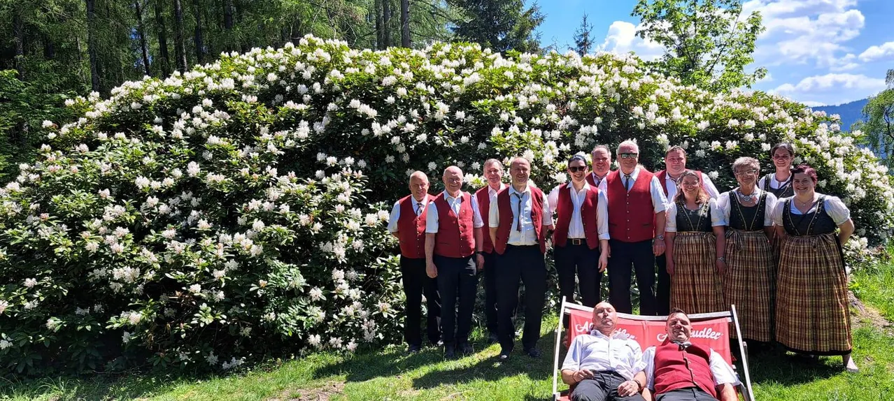 Eine Gruppe von Männern in roten Westen posiert für ein Foto vor einem weißen Blumenbusch. Ein Mann sitzt auf einem roten Stuhl im Vordergrund.