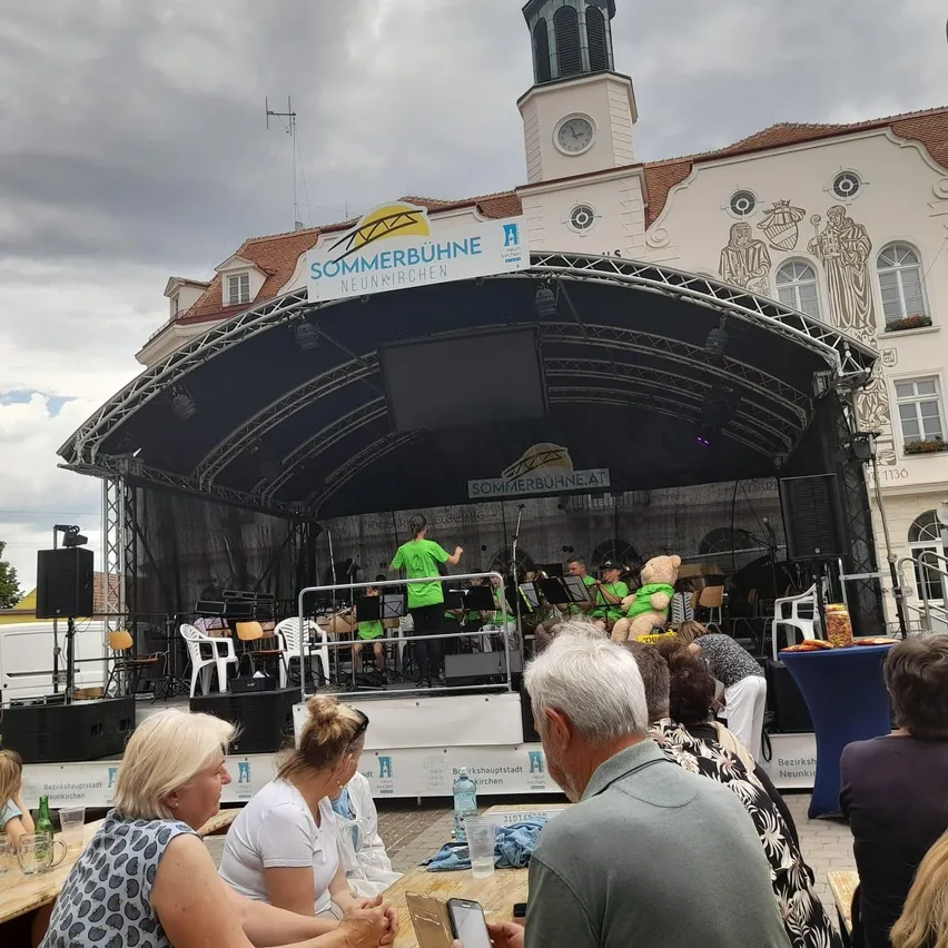 Ein Konzert auf der Sommerbühne Neunkirchen mit Musikern auf der Bühne und einem zuschauenden Publikum. Die Bühne hat eine große Leinwand und einen schwarzen Hintergrund.