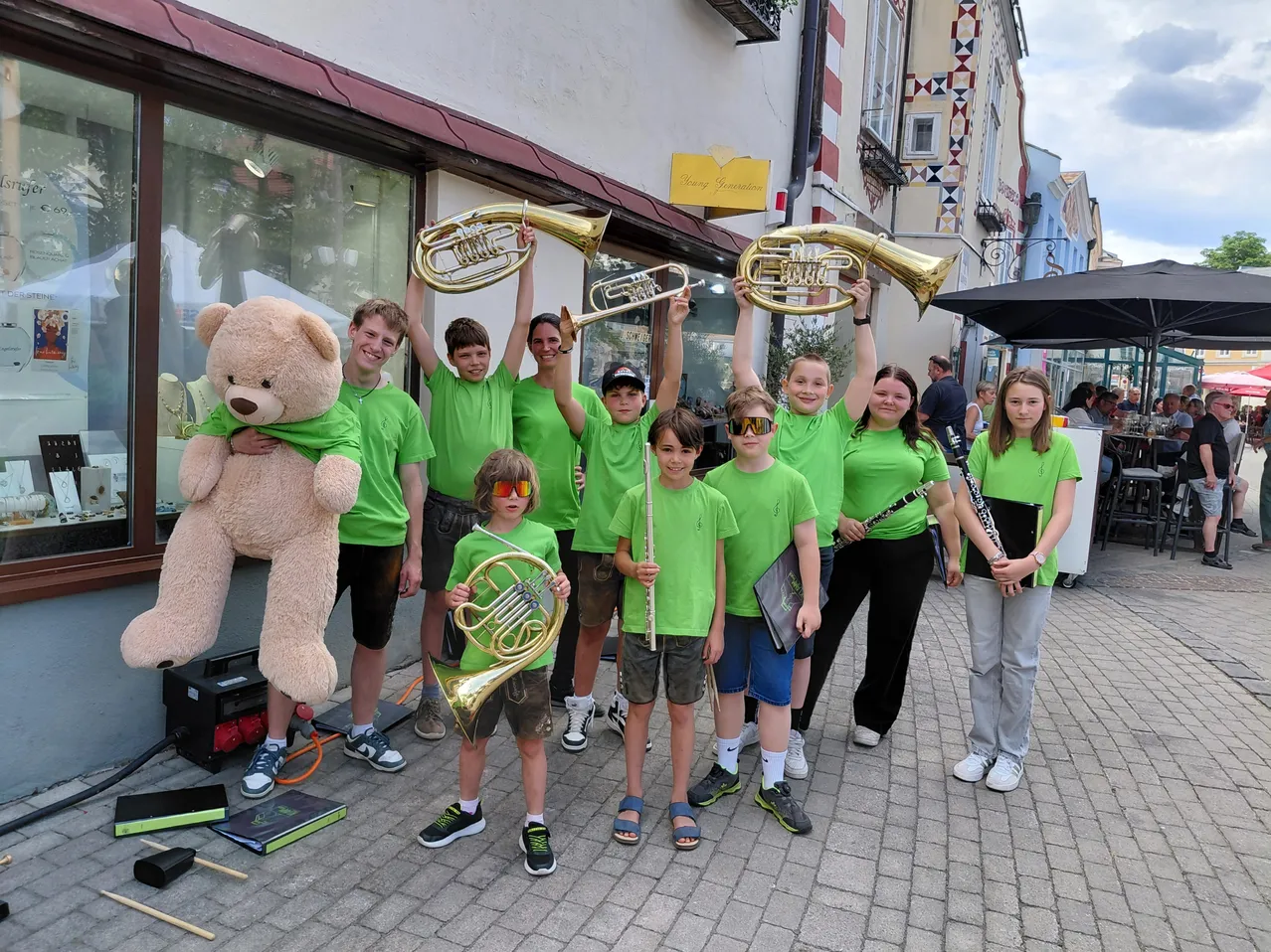 Eine Gruppe junger Musiker in grünen T-Shirts posiert mit ihren Instrumenten vor einem Gebäude, wobei eine Person einen Teddybären hält.