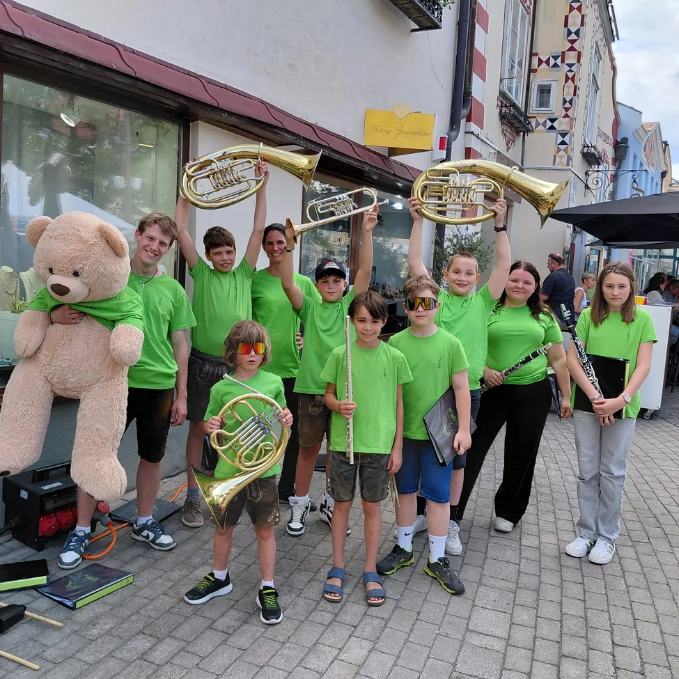 Eine Gruppe junger Musiker in grünen T-Shirts posiert mit ihren Instrumenten vor einem Gebäude, wobei eine Person einen Teddybären hält.