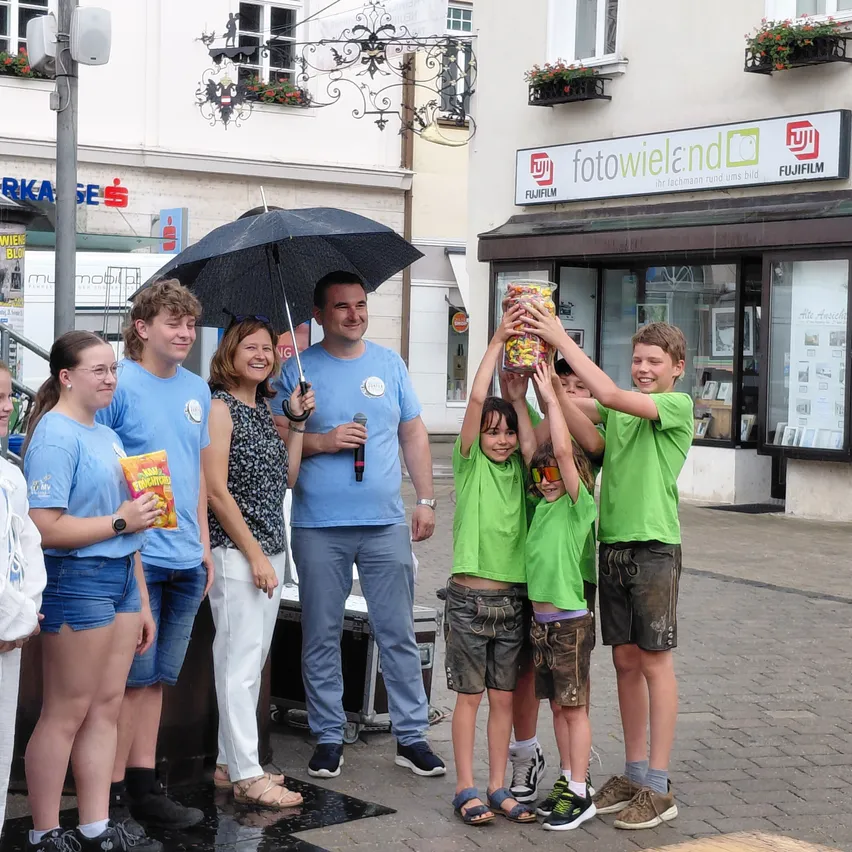 Eine Gruppe von Menschen steht im Regen und hält eine Tüte Popcorn hoch. Dahinter befindet sich ein Geschäft mit einem Fujifilm-Schild.