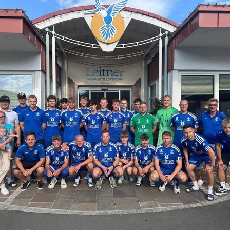 Eine Gruppe von Fußballspielern in blauen Trikots, einige in Grün, posiert für ein Foto vor dem Leitner Thermalhotel Loipersdorf.