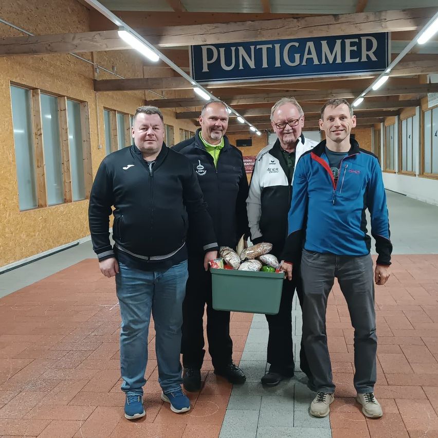 Vier Männer stehen in einem Innenflur, zwei halten einen grünen Container mit Lebensmitteln. Ein Schild mit der Aufschrift 'PUNTIGAMER' ist über ihnen.